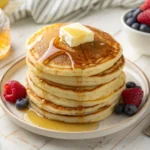 Fluffy stack of pancakes with melting butter, syrup drizzle, and fresh berries on a wooden kitchen counter.