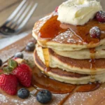 Fluffy pancakes stacked high with syrup, butter, and fresh berries on a rustic wooden kitchen counter.