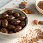 Close-up of shiny chocolate-covered almonds in a small bowl, with whole almonds and cocoa powder surrounding them.