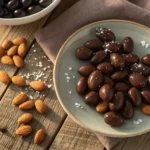 Plate of chocolate-covered almonds scattered on a rustic wooden table with soft natural lighting.