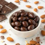 Close-up of chocolate-covered almonds in a bowl with scattered almonds and chocolate pieces